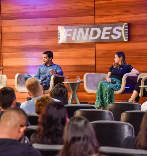 Sala cheia de ouvintes, com 4 pessoas em cima de um palco discursando sobre inovação