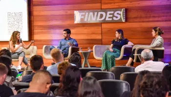 Sala cheia de ouvintes, com 4 pessoas em cima de um palco discursando sobre inovação